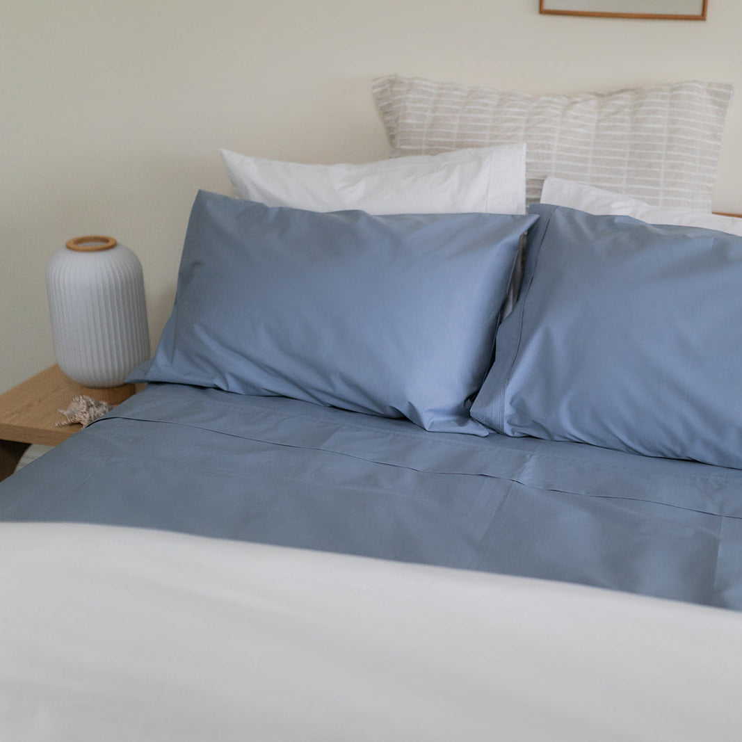 Blue sheet set on a bed with contrasting white pillows and nightstand with  white lamp