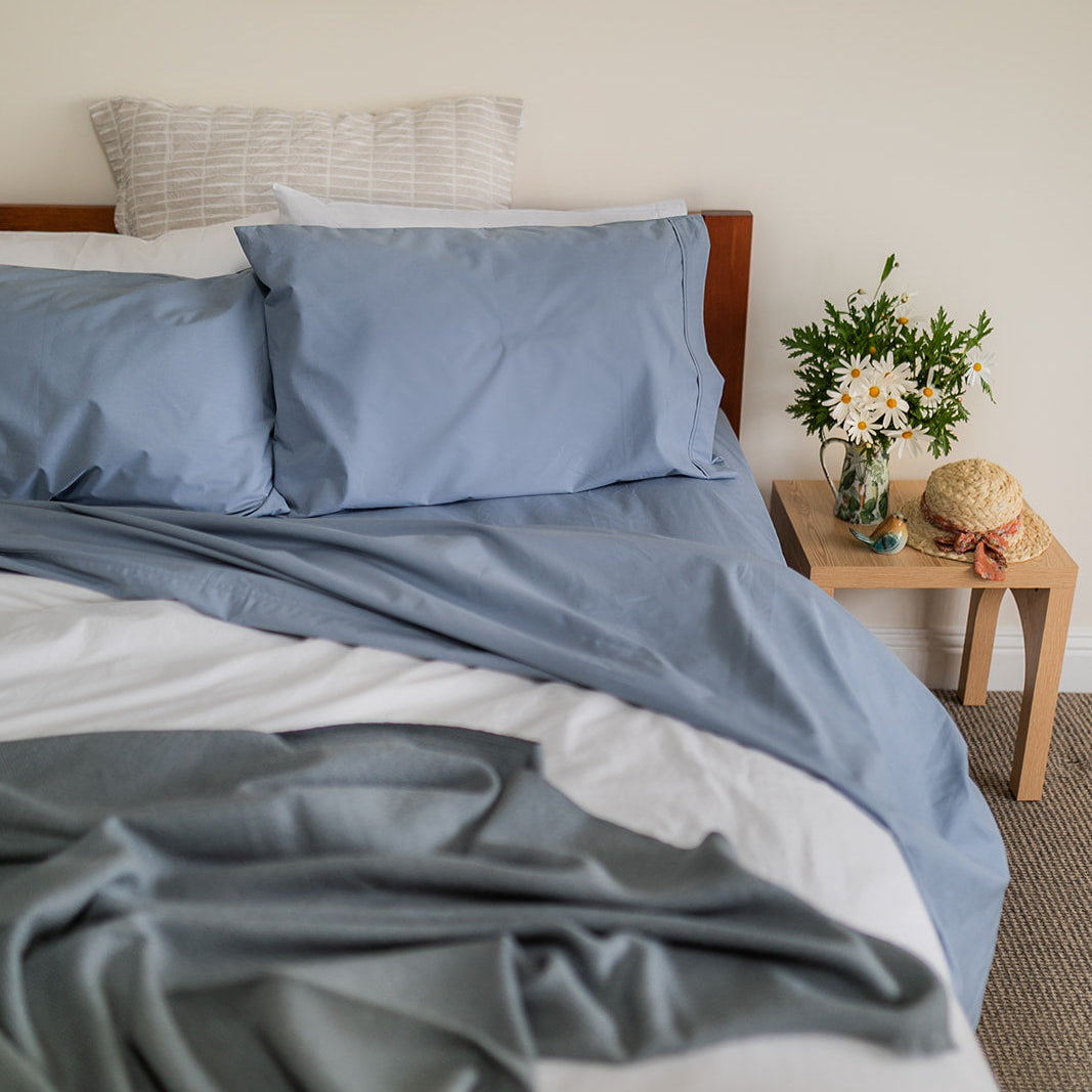 Bed with organic cotton blue sheet set, next to a side-table with flowers & straw hat