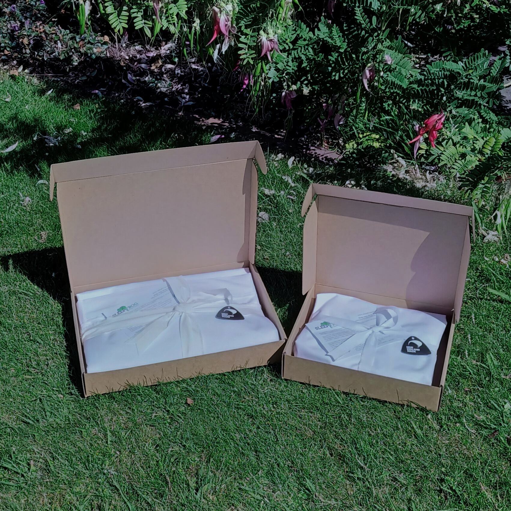 Open cardboard gift-box style packages containing white cot sheets with a made-in -NZ logo , placed on a grassy surface with Kakabeak flowers in the background.