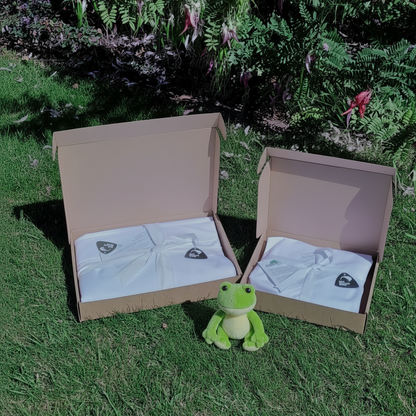 Open cardboard gift-box with white cot sheets, showing a made-in -NZ tag, placed in a garden area with a  soft green frog toy. 